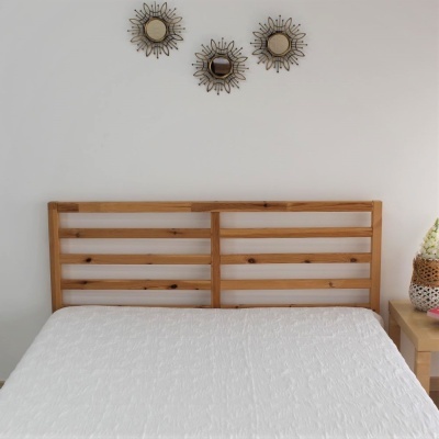 Cabeceira de cama de madeira clara com ripas horizontais, colcha branca, três espelhos decorativos na parede branca, mesa de cabeceira com vaso