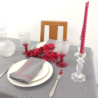Mesa posta com toalha xadrez, pratos brancos, copos vermelhos e castiçal de vidro com vela vermelha