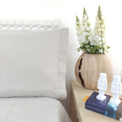 Cama com cabeceira cinza, vaso de madeira com flores brancas numa mesa com livros e objetos decorativos brancos