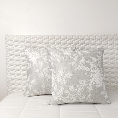 Duas almofadas quadradas cinzentas com padrão floral branco sobre cama branca com cabeceira acolchoada.