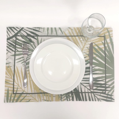 Jogo americano com padrão de folhas verde e dourado, prato branco, talheres de metal e copo transparente