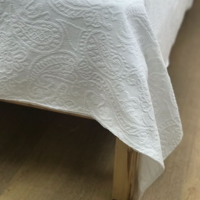 Toalha ou colcha branca com padrão em relevo cobrindo cama de madeira.