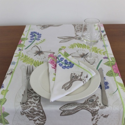 Mesa posta com toalha, guardanapo com padrão de coelhos e flores, prato branco, talheres e copo transparente