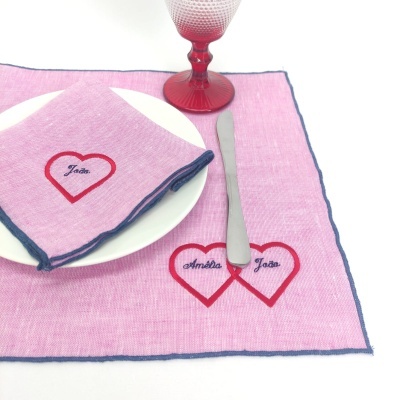 Conjunto de mesa com guardanapo cor-de-rosa bordado com coração e nomes, prato branco, faca e copo vermelho.