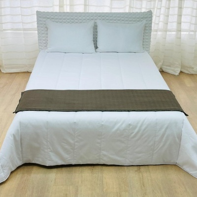 Cama de casal com edredão branco e faixa castanha numa sala com chão de madeira e cortinas brancas