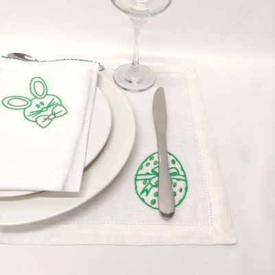 Mesa posta com pratos brancos, guardanapo com coelho bordado e espaço americano com padrão verde e talheres.