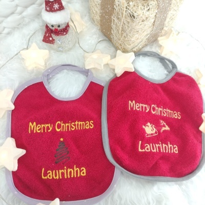 Dois babetes vermelhos bordados com texto de Natal, rodeados por luzes em forma de estrela e decoração de Natal.