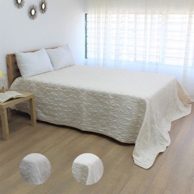 Cama com colcha branca texturizada e almofadas, mesa de madeira e cortinas brancas