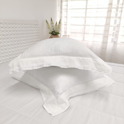 Conjunto de almofadas brancas com bordas bordadas sobre cama branca, em quarto iluminado pela janela.