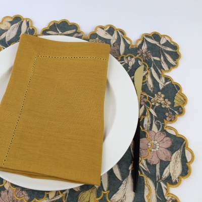 Conjunto de mesa com prato branco, guardanapo amarelo mostarda, toalha floral com bordas onduladas e talheres dourados e pretos.
