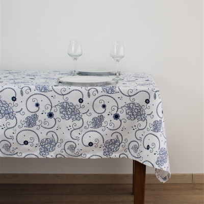 Toalha de mesa branca com padrão azul floral e arabescos sobre mesa de madeira com dois copos e um prato de vidro