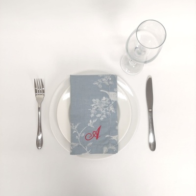 Mesa posta com prato branco, guardanapo azul floral com letra A vermelha, talheres e copo