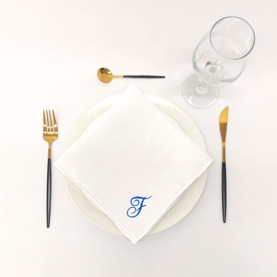 Conjunto de louça com prato branco, talheres dourados e pretos, guardanapo branco com letra 'F' azul e copo de vidro