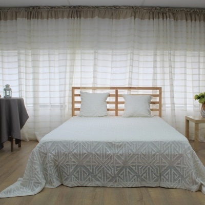 Quarto com cama de madeira clara, colcha branca com padrão cinzento, duas almofadas brancas, mesas laterais em madeira clara, cortinas brancas e chão em madeira clara