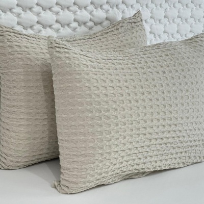 Duas almofadas bege com padrão texturizado, sobre superfície branca com pano branco texturizado ao fundo.