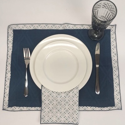 Conjunto de jantar com pratos brancos, talheres e copo em toalha azul escura com padrões