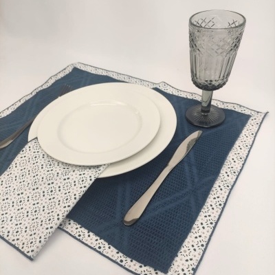Conjunto de mesa com pratos brancos, guardanapo, talheres metálicos, copo cinza e suporte azul escuro