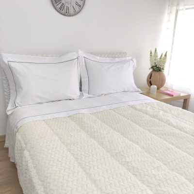 Cama com colcha branca, almofadas brancas, relógio de parede e mesa de cabeceira com vaso.