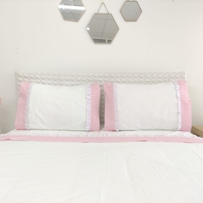 Cama com roupa de cama branca e rosa com almofadas e lençol, frente a parede branca com três espelhos hexagonais