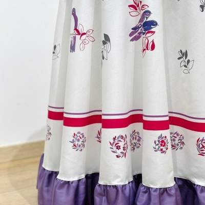 Tecido branco com motivos de folhas e pássaros em várias cores, borda vermelha e volado roxo.