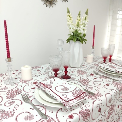 Mesa posta com toalha branca e padrão vermelho, copos e castiçais, jarro com flores brancas