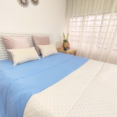 Cama com edredão azul claro e lençol branco com padrão de pontos azuis junto a mesa de cabeceira com vaso e telefone