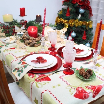 Mesa decorada com toalha de Natal, pratos brancos e vermelhos, copos vermelhos, velas e pinhas, árvore de Natal ao fundo