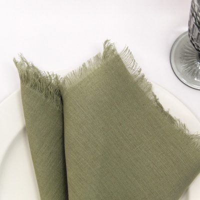 Guardanapo verde-oliva de tecido com bordas desfiadas junto a prato branco e vidro faceteado
