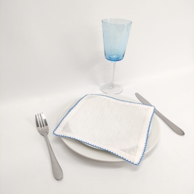 Prato branco com guardanapo branco com borda azul, talheres em aço inox e copo alto azul.