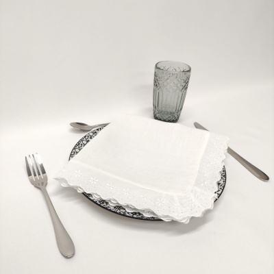Conjunto de mesa com prato decorativo preto e branco, guardanapo branco bordado, talheres em aço inox e copo cinza translúcido.