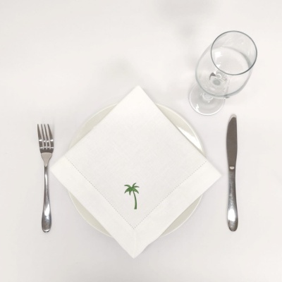 Conjunto de jantar com prato branco, guardanapo com bordado de palmeira, talheres e copo.