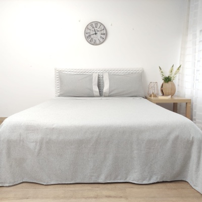 Cama de casal com colcha e almofadas cinzentas, cabeceira branca, relógio de parede e mesa de cabeceira com vaso e flores.