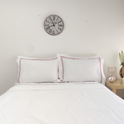 Cama com roupa branca, relógio de parede com números romanos e mesa de cabeceira com relógio e vaso de flores