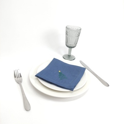 Conjunto de louça branca, guardanapo azul bordado com árvore de Natal, talheres metálicos e copo de vidro texturizado