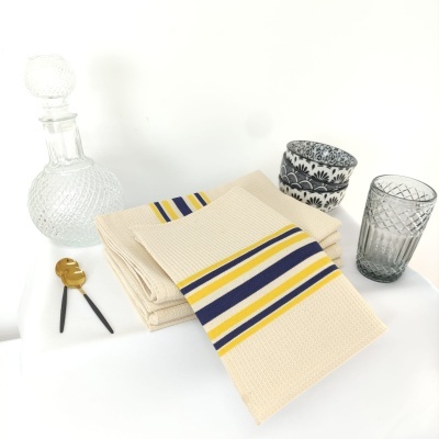 Toalhas bege com riscas coloridas, garrafa de vidro e copos decorativos sobre mesa branca