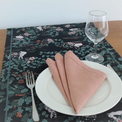 Mesa posta com prato branco, guardanapo rosa, garfo e copo em toalha com padrão floral escuro.