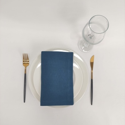 Conjunto de jantar com prato branco, guardanapo azul, talheres preto e dourado, copo transparente em fundo branco.