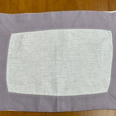 Individual de mesa retangular em tecido branco e lilás sobre superfície de madeira.