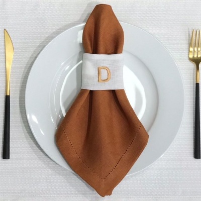 Guardanapo castanho com anel branco e letra D em prato branco com talheres dourados e pretos sobre toalha branca