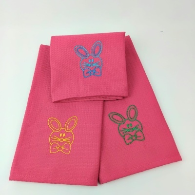 Três panos de cozinha rosa com bordados coloridos de coelhos.