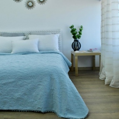 Quarto com cama azul claro, mesa de cabeceira, vaso preto e cortinas brancas