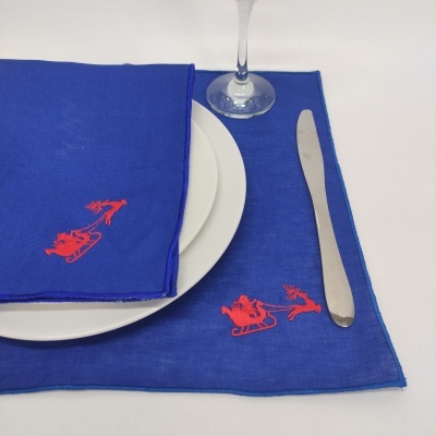 Guardanapo e individual azul com bordado de Pai Natal em trenó vermelho, prato branco, talher e copo.