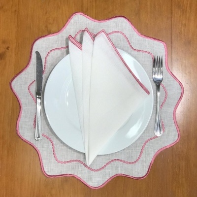 Conjunto de mesa com prato branco, guardanapo branco com borda rosa, sousplat bege com borda rosa, e talheres prateados sobre mesa de madeira