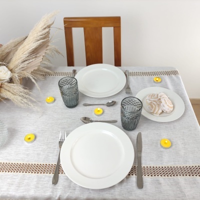 Mesa posta com pratos brancos, copos de vidro cinza, talheres de aço, velas amarelas e biscoitos numa toalha cinza e castanha.