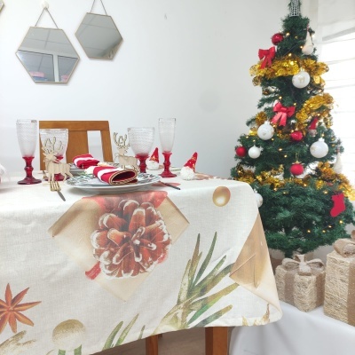 Mesa de jantar com toalha natalícia, pratos, copos e talheres, de frente para árvore de Natal decorada com presentes.