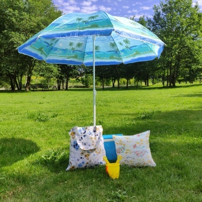 Guarda-sol azul com padrão de praia num parque junto a almofadas e balde amarelo