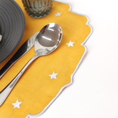 Talheres de metal sobre tecido amarelo com estrelas brancas e borda prateada, ao lado um copo de vidro e um prato cinza