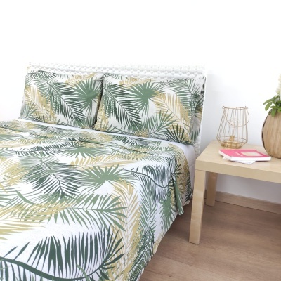 Roupa de cama com padrão de folhas tropicais verdes e amarelas em cama branca junto a mesa de madeira