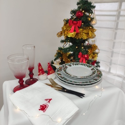 Conjunto de pratos, copos, talheres e guardanapo com decoração natalícia numa mesa ao lado de uma árvore de Natal decorada.