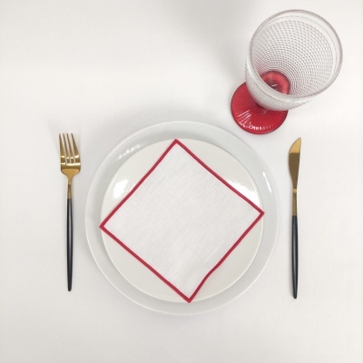 Conjunto de mesa com prato branco, guardanapo com borda vermelha, talheres com cabo preto e dourado, e copo transparente com suporte vermelho.